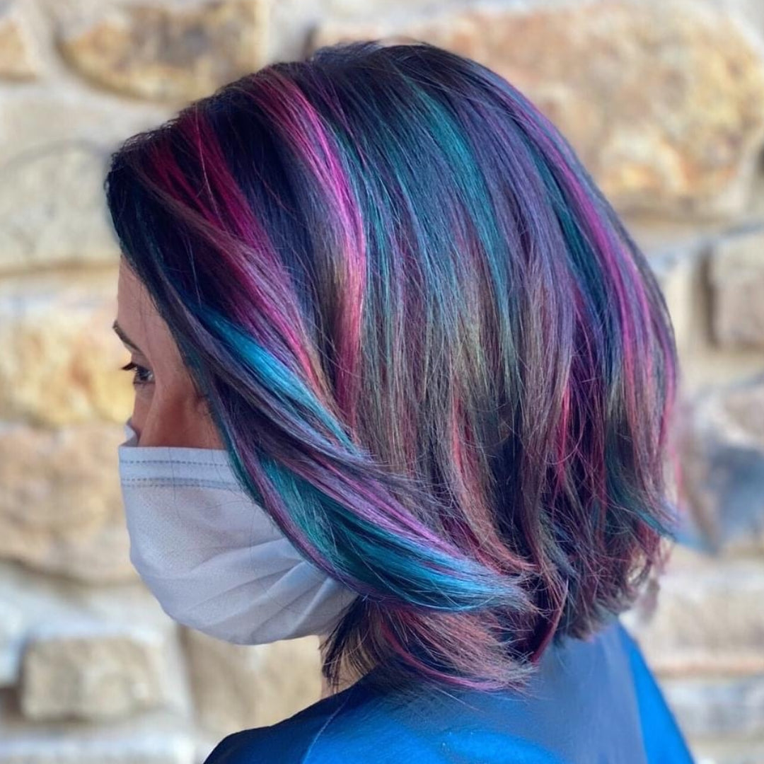 This is an image of an Ippodaro Natural Salon customer who just received a series of highlights featuring a really pretty blue and pink intermingled with the rest of her brown hair. We are looking at her profile as she stands in front of a rock wall, wearing a mask.